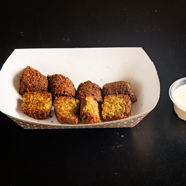 A white paper tray filled with golden-brown falafel pieces, some revealing green interiors, with a side of sauce.