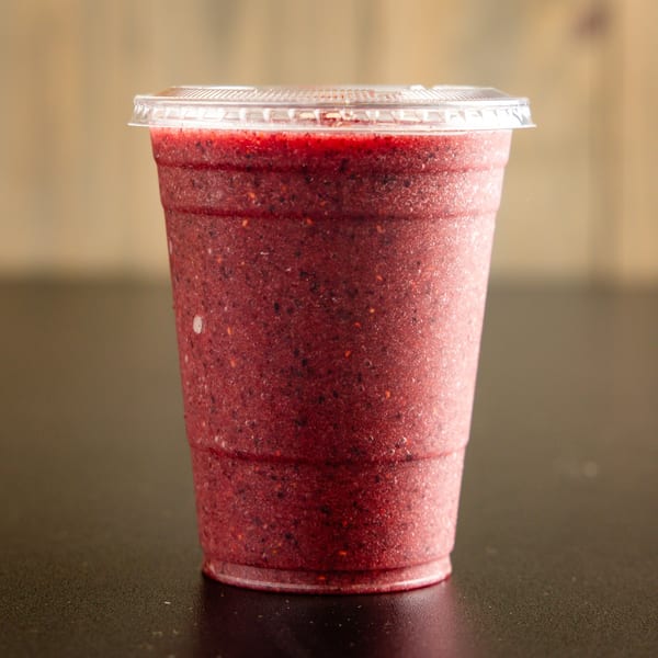 A vibrant red berry smoothie in a clear plastic cup with a lid, on a dark table.