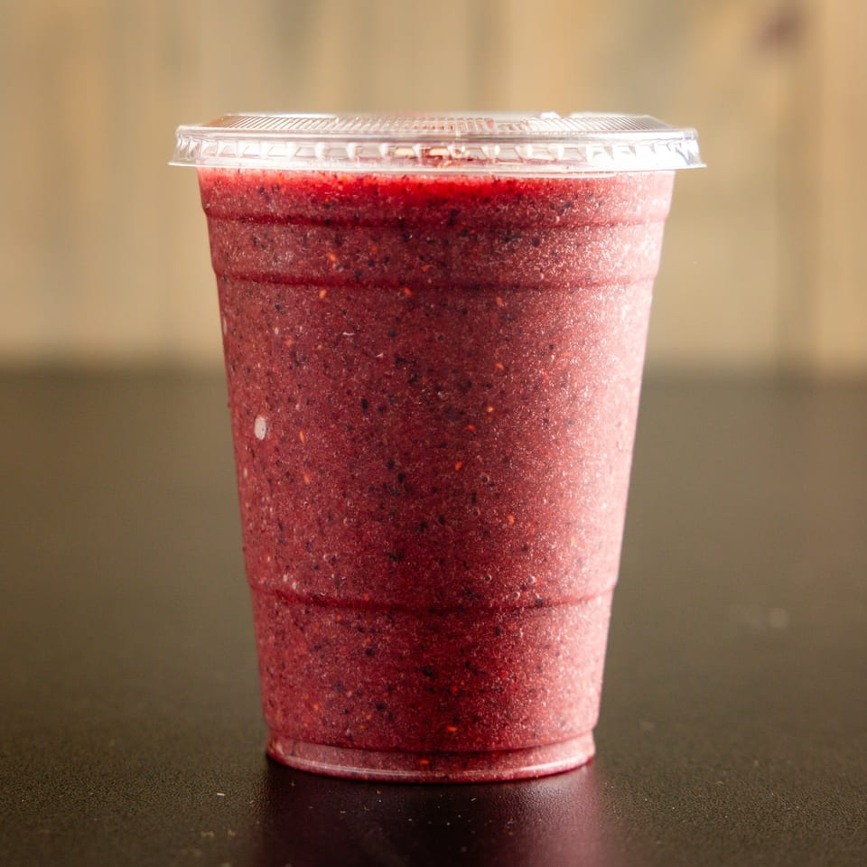 A vibrant red berry smoothie in a clear plastic cup with a lid, on a dark table.